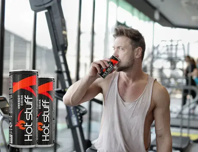 A fit man who drinks Hot Stuff Energy Drink in a gym, with workout equipment and large windows in the background.